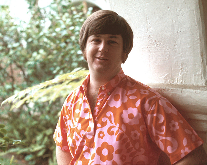 Beach Boys co-founder and musical genius wearing floral shirt, standing outdoors near white wall and greenery.