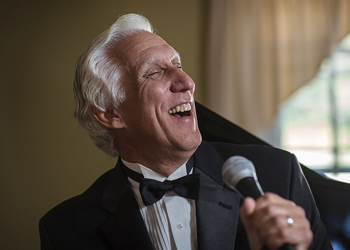 Older man in tuxedo laughing joyfully, holding a microphone, capturing a moment of unexpected wedding drama.