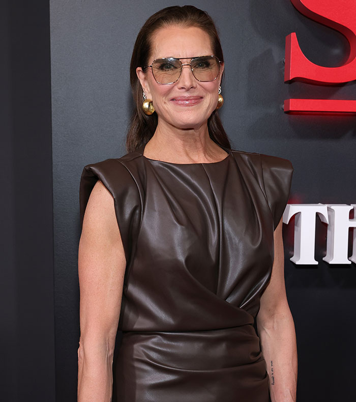 Brooke Shields wearing sunglasses and a brown leather dress at a red carpet event about Meghan Markle podcast interview.