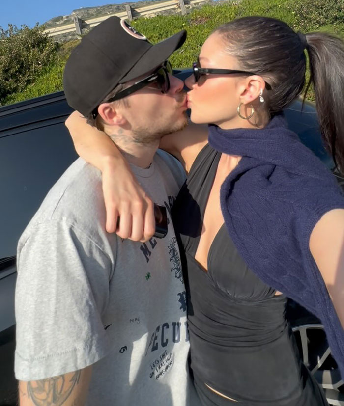 Brooklyn Beckham kissing woman outdoors, wearing sunglasses and casual clothing amid alleged no contact feud with David and Victoria Beckham.