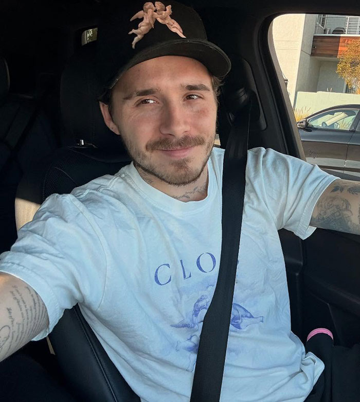Brooklyn Beckham wearing a black cap and white shirt, sitting in a car with a calm expression amid family feud.