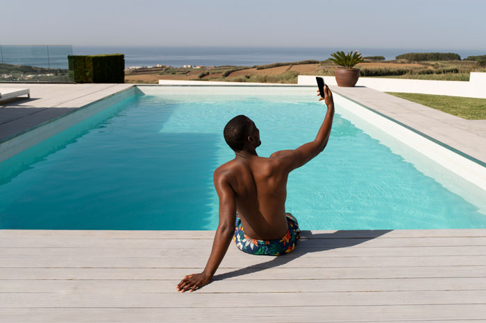 Man sitting by pool taking selfie, illustrating man calling cops after bro and family broke into his home to use pool.