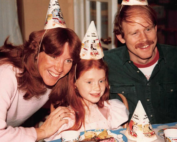 Family celebrating a birthday with Bryce Dallas Howard as a child wearing party hats in a vintage photo capturing her childhood memory.