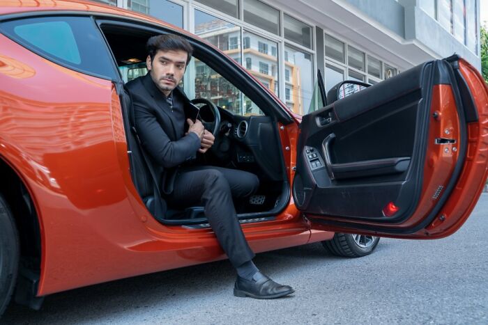 Man in a black suit seated in an orange sports car, showcasing out-of-touch rich people flaunting their wealth outdoors.