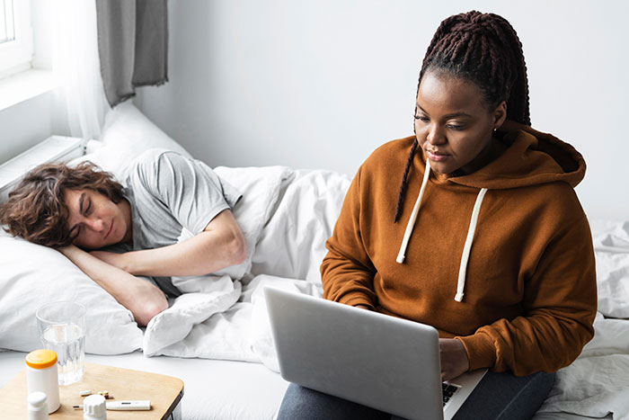 Teenage boy resting in bed after accident while woman in hoodie works on laptop nearby in bright room Teenage boy resting in bed after accident while woman in hoodie works on laptop nearby in bright room