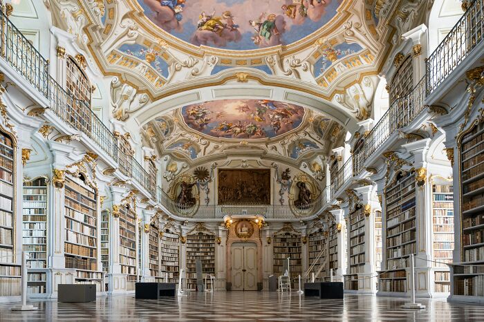 Ornate historic library interior with towering bookshelves, intricate ceiling frescoes, and elegant architectural details.