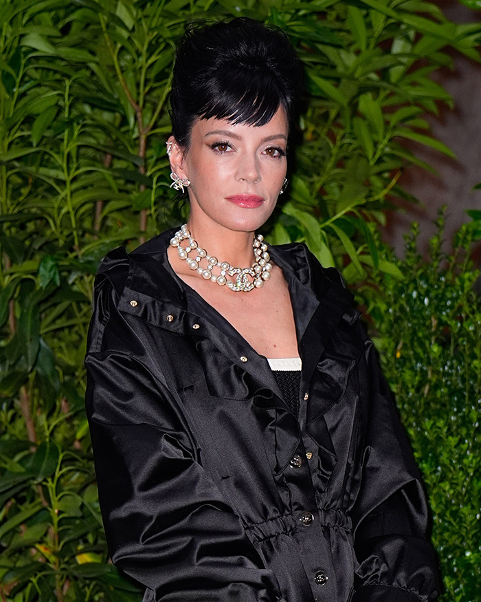 Woman with dark hair styled up wearing a black jacket and pearl necklace standing in front of green plants, celebrities with body abnormalities.