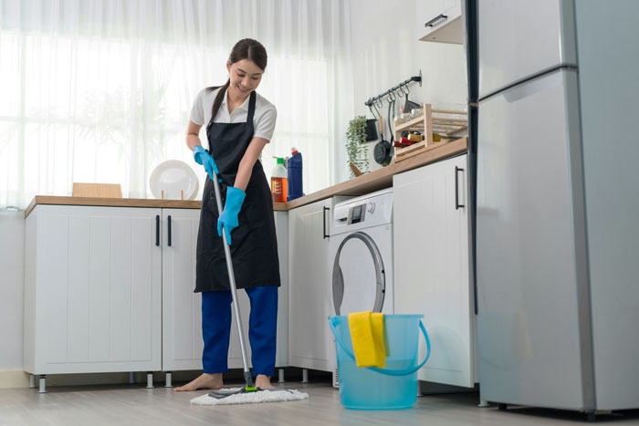 Woman cleaning kitchen floor with mop and gloves, illustrating venue host sending $1200 cleaning bill after guest stay dispute.