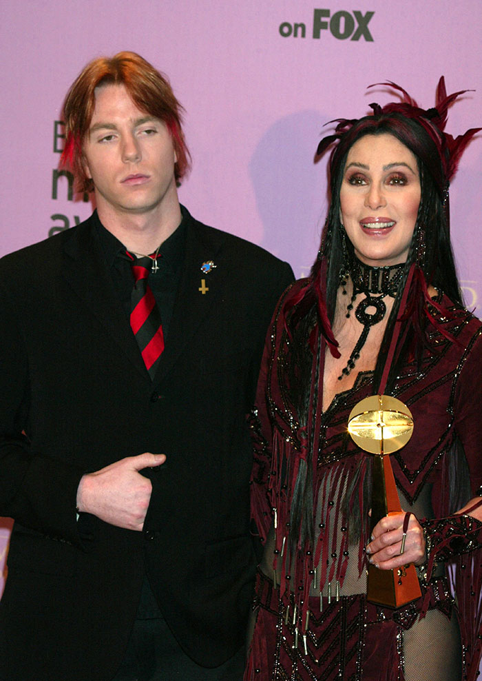 Cher in a red and black outfit holding an award, standing next to her son amid alleged kidnap and overdose news.