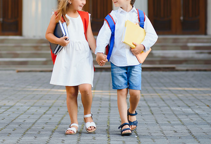 Two 5th grade children with backpacks walking hand in hand outside school, holding books and smiling at each other. Two 5th grade children with backpacks walking hand in hand outside school, holding books and smiling at each other.