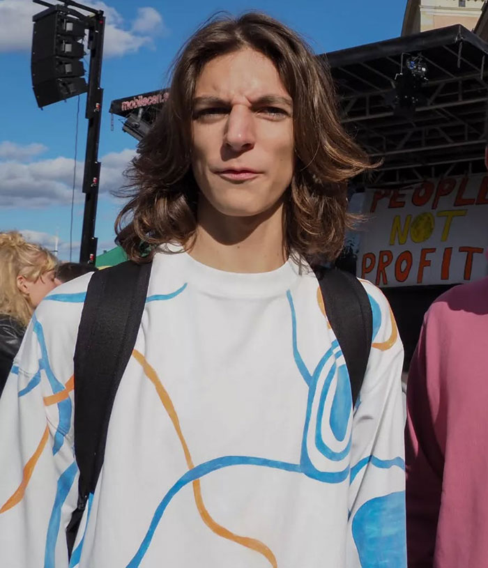 Young man with long hair wearing a colorful shirt and black backpack at an outdoor event about people and profit.