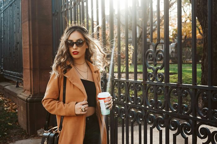 Stylish woman in sunglasses holding coffee, standing by iron gate outdoors, representing hobbies that give netizens the ick.