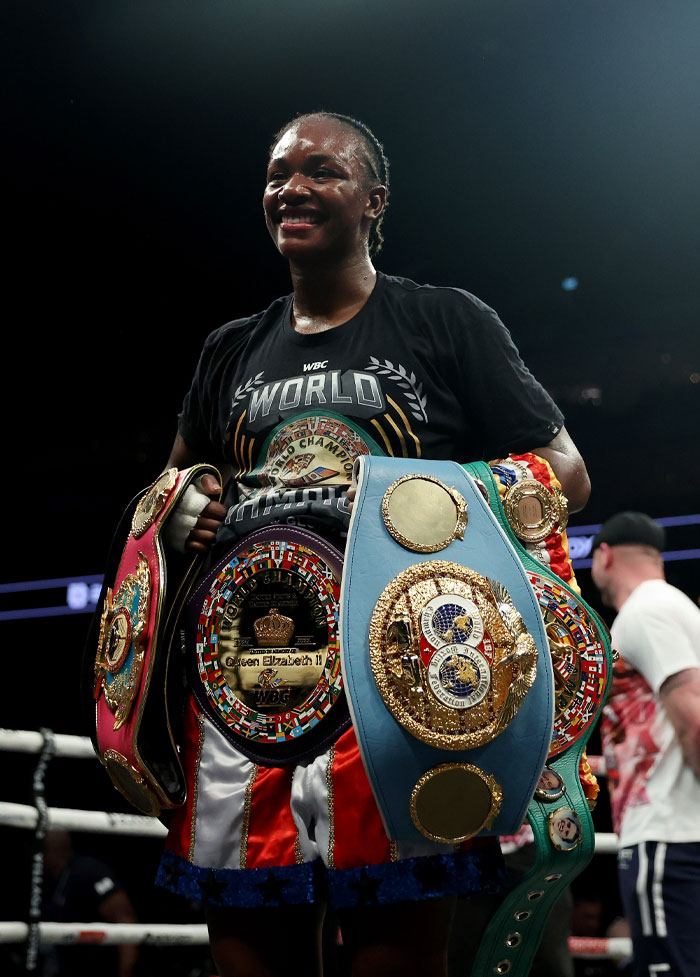 Claressa Shields smiling in the ring with multiple championship belts showcasing her boxing champ status and net worth.