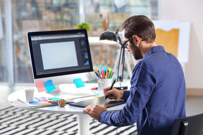 Designer working on digital tablet at desk with computer, focusing on client approvals and design follow-ups. Designer working on digital tablet at desk with computer, focusing on client approvals and design follow-ups.
