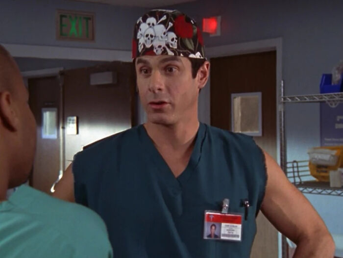 Male former Hollywood star dressed as a doctor in scrubs and skull cap, representing a normal career transition.