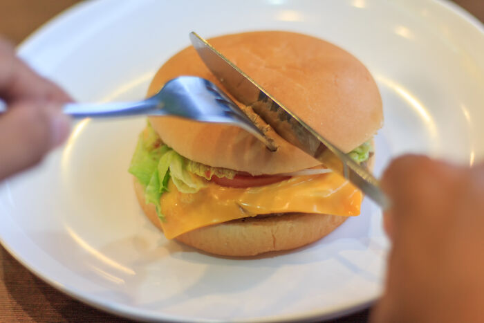 Person cutting a cheeseburger with knife and fork, illustrating things non-Americans do like breaking spaghetti for Italians.