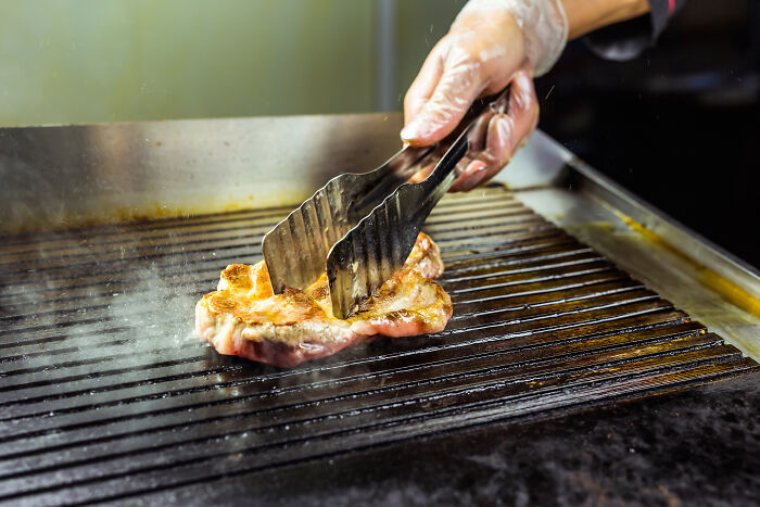 Grilled meat being cooked with tongs on a hot grill, illustrating Americans reveal things non-Americans do.