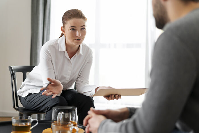 Female therapist in a white shirt holding a clipboard, talking to a client during a counseling session in a bright room.