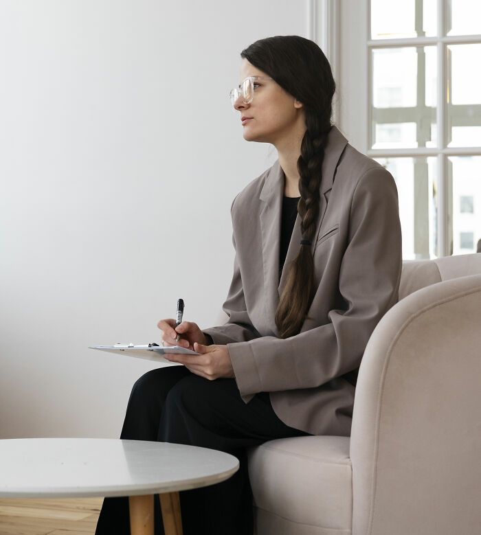 Therapist with glasses and braided hair holding a pen and clipboard, reflecting during a counseling session in a bright room.