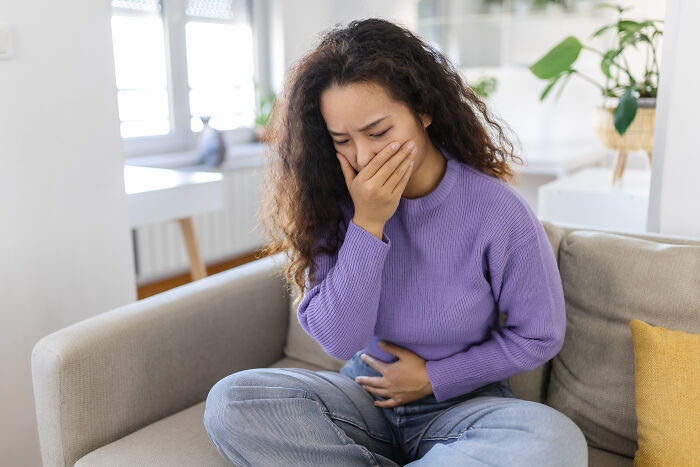 Woman sitting on a couch looking distressed, holding her stomach and covering her mouth, illustrating postpartum OCD symptoms.