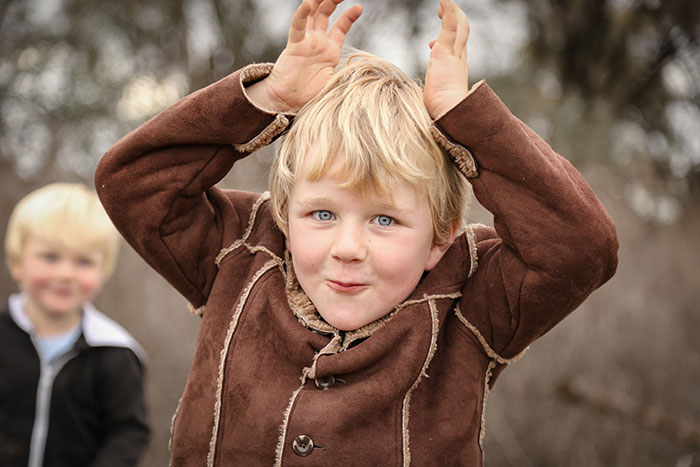 Young blond child in brown coat making playful gestures outdoors, relating to babysitter refusing one specific child situation.