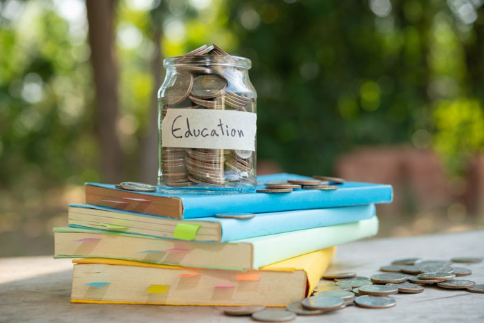 Glass jar labeled education filled with coins on colorful stacked books outdoors representing saving money.