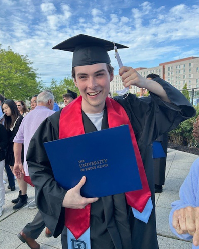 College student celebrating graduation outdoors, holding diploma in cap and gown with red stole on a sunny day.
