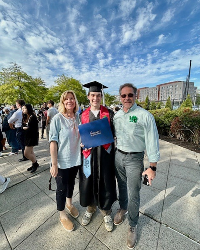 College student in graduation gown holding diploma, smiling with family outdoors on a sunny day after ceremony.