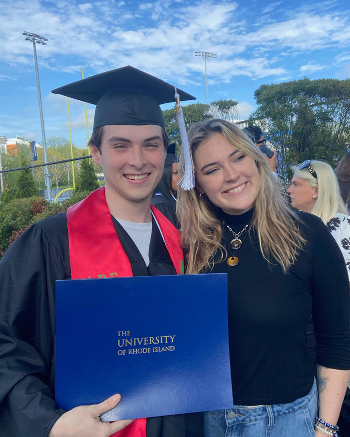 College student in graduation gown holding diploma, smiling with a friend outdoors under a clear blue sky.