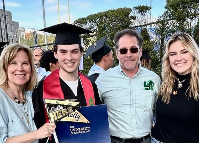 College student in graduation cap and gown holding diploma with family celebrating at outdoor ceremony.