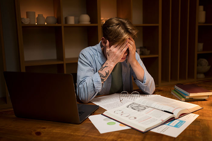 Stressed woman studying at a desk with open books and laptop, facing risk of not graduating over shared thesis issues
