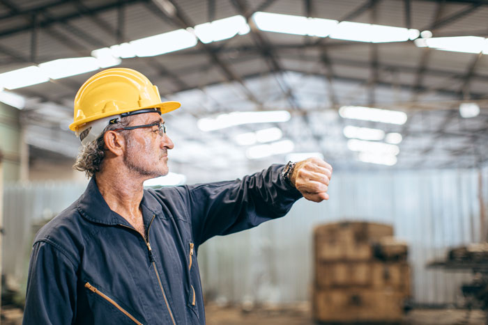 Middle-aged factory worker in yellow helmet checking time, illustrating company penny-pinching backfiring as workers stop extra effort. Middle-aged factory worker in yellow helmet checking time, illustrating company penny-pinching backfiring as workers stop extra effort.