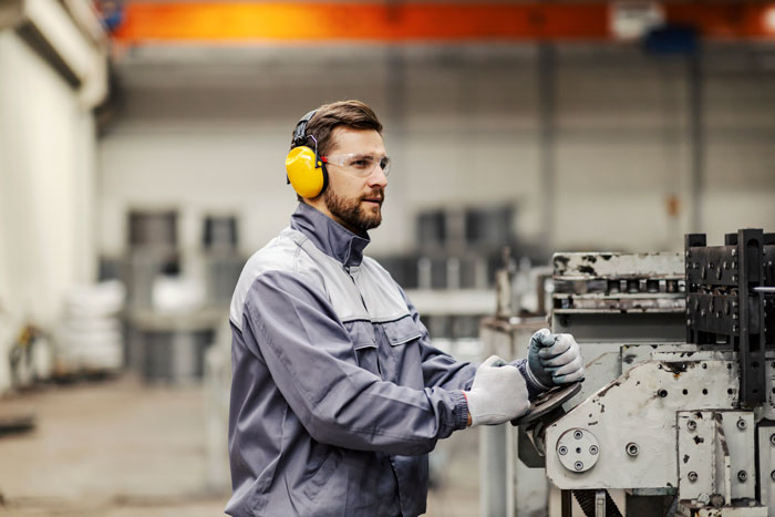 Factory worker wearing safety gear operating machinery, illustrating company penny-pinching backfiring with reduced employee effort. Factory worker wearing safety gear operating machinery, illustrating company penny-pinching backfiring with reduced employee effort.