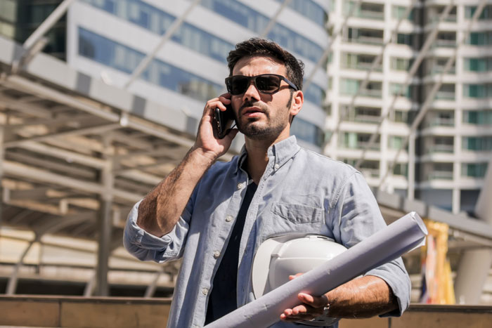 Man speaking loudly on phone outdoors wearing sunglasses, holding construction helmet and blueprints in urban setting.