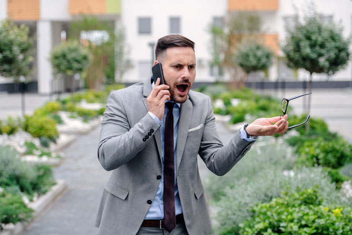 Man in gray suit speaking loudly on phone outdoors, displaying frustration, fitting habitually speaking loudly in the library behavior.
