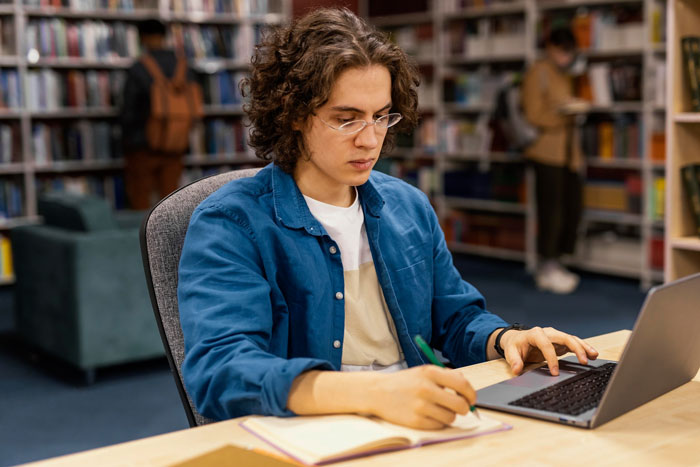 Young man studying in the library, working on laptop and notebook, while local regulars observe the loud behavior.