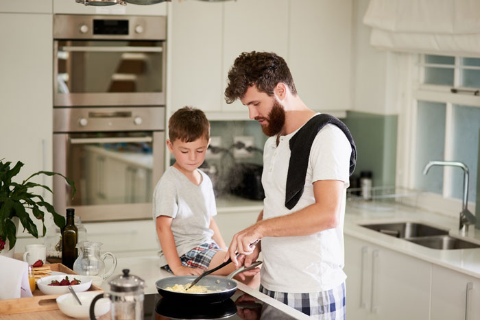 Man cooking breakfast for kids in modern kitchen while young boy watches, highlighting playing chef for her kids concept.