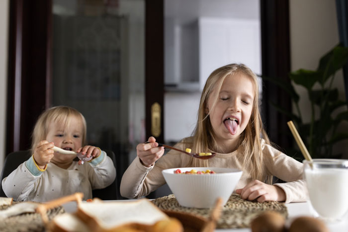 Two children eating breakfast cereal at a table while their uncle plays chef for them in a home kitchen.