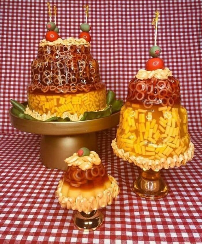 Layered gelatin mold with macaroni and cheese, topped with olives and cherry tomatoes on checkered tablecloth, gross foods.