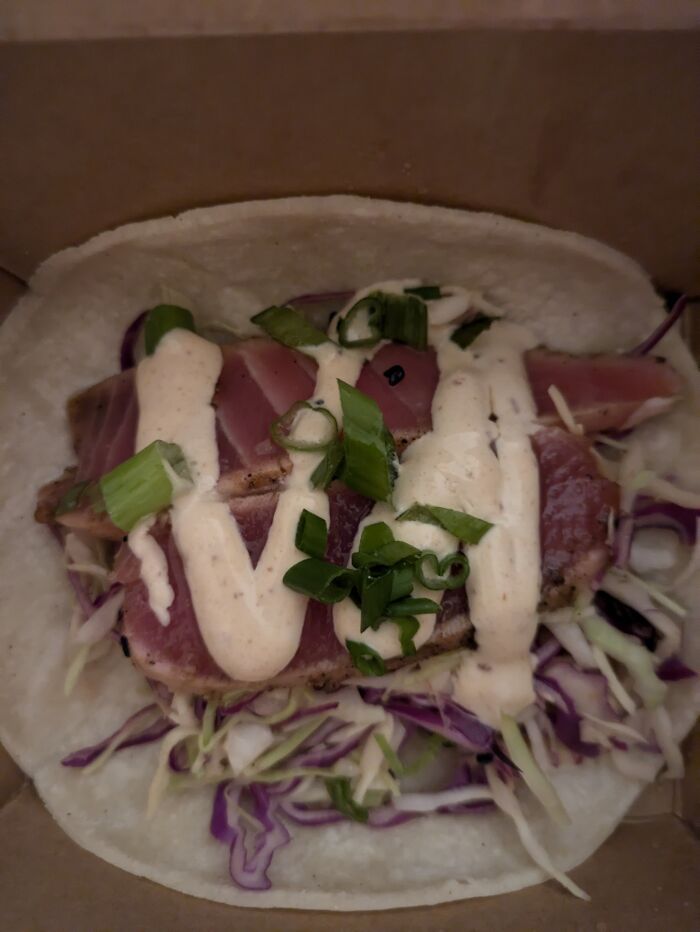 Seared fish taco topped with creamy sauce and green onions on a bed of shredded cabbage and tortilla, unusual gross foods.