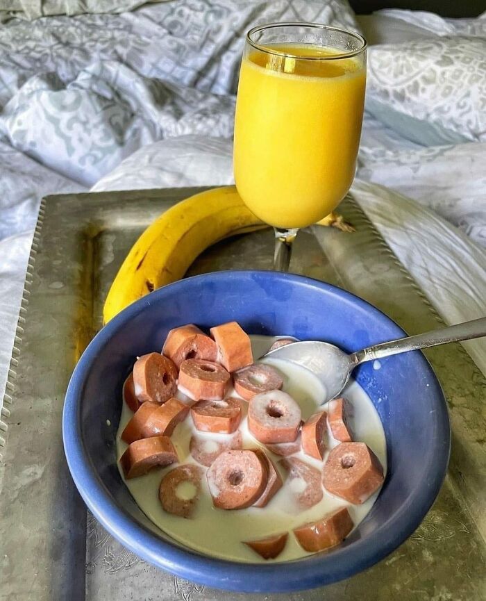 Unusual combination of sliced hot dogs in milk in a bowl with a banana and a glass of orange juice on a tray.