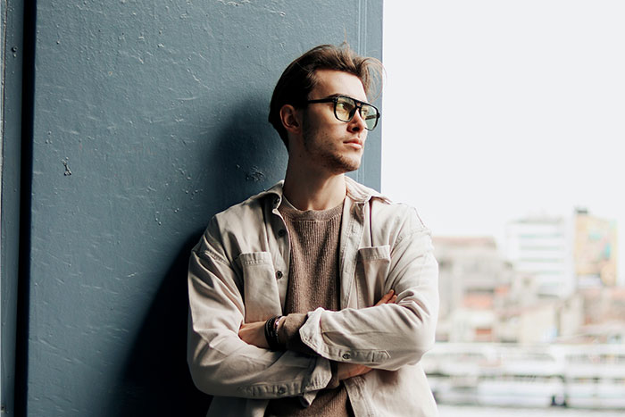 Young man wearing glasses and a beige jacket, standing with arms crossed, reflecting on life after peaking in high school.