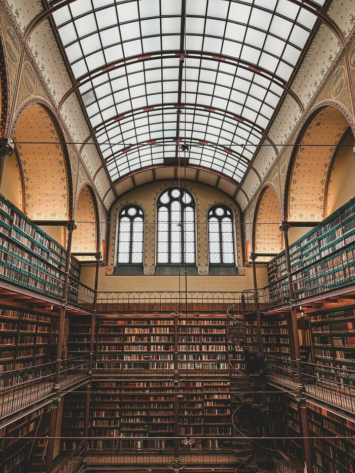 Grandiose historic library interior with tall bookshelves, ornate arches, and large arched windows under a glass ceiling.