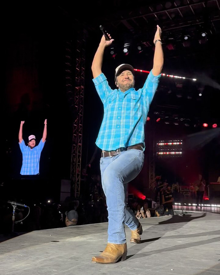 Country star performing on stage in a blue shirt and jeans, raising arms during a live concert event.