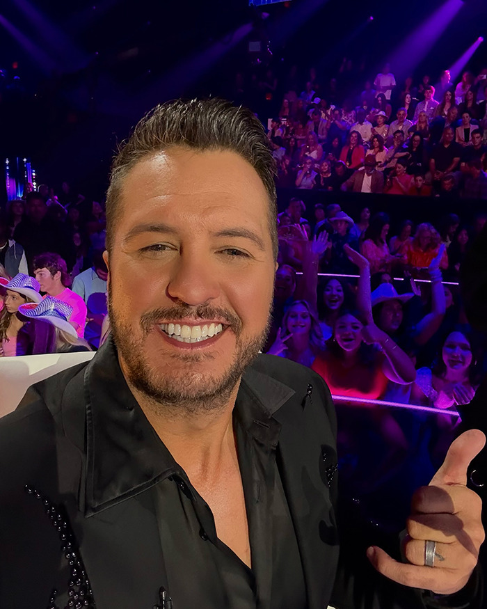 Country star smiling and giving thumbs up during a live concert with a cheering crowd in the background.