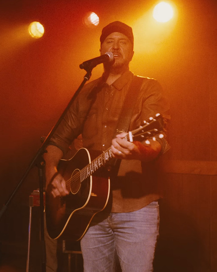 Country star performing on stage with guitar, wearing a cap and brown shirt under warm concert lighting.