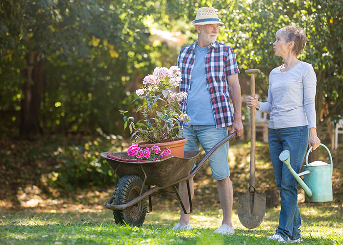 Middle-aged couple gardening outdoors, woman holding watering can and man with wheelbarrow, illustrating garden landscaping.