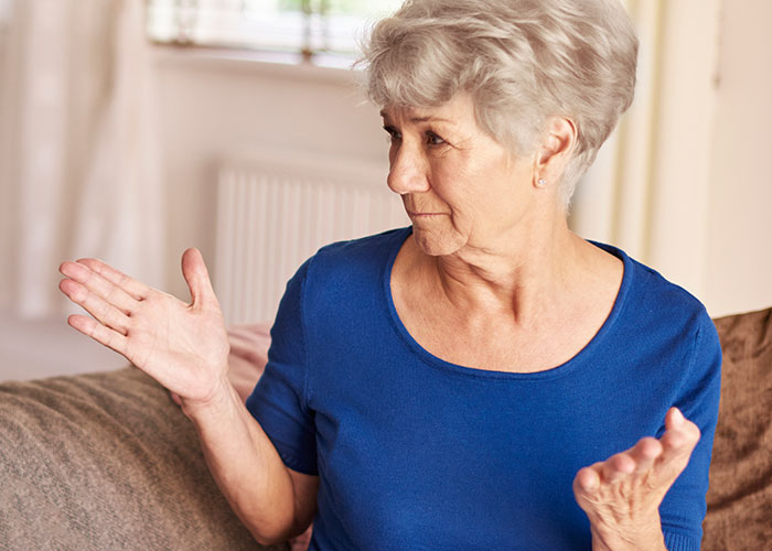 Older woman in blue shirt gesturing with hands while sitting indoors, representing MIL landscaping garden without permission.