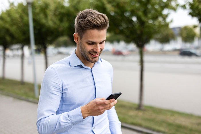 Young man in blue shirt smiling while checking his phone outdoors near trees and a street in the background