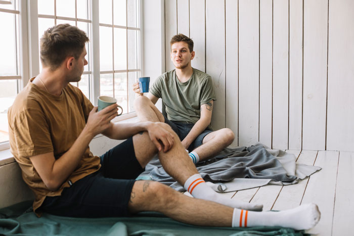 Two young men casually sitting and talking by the window, sharing a conversation about a job opportunity found.
